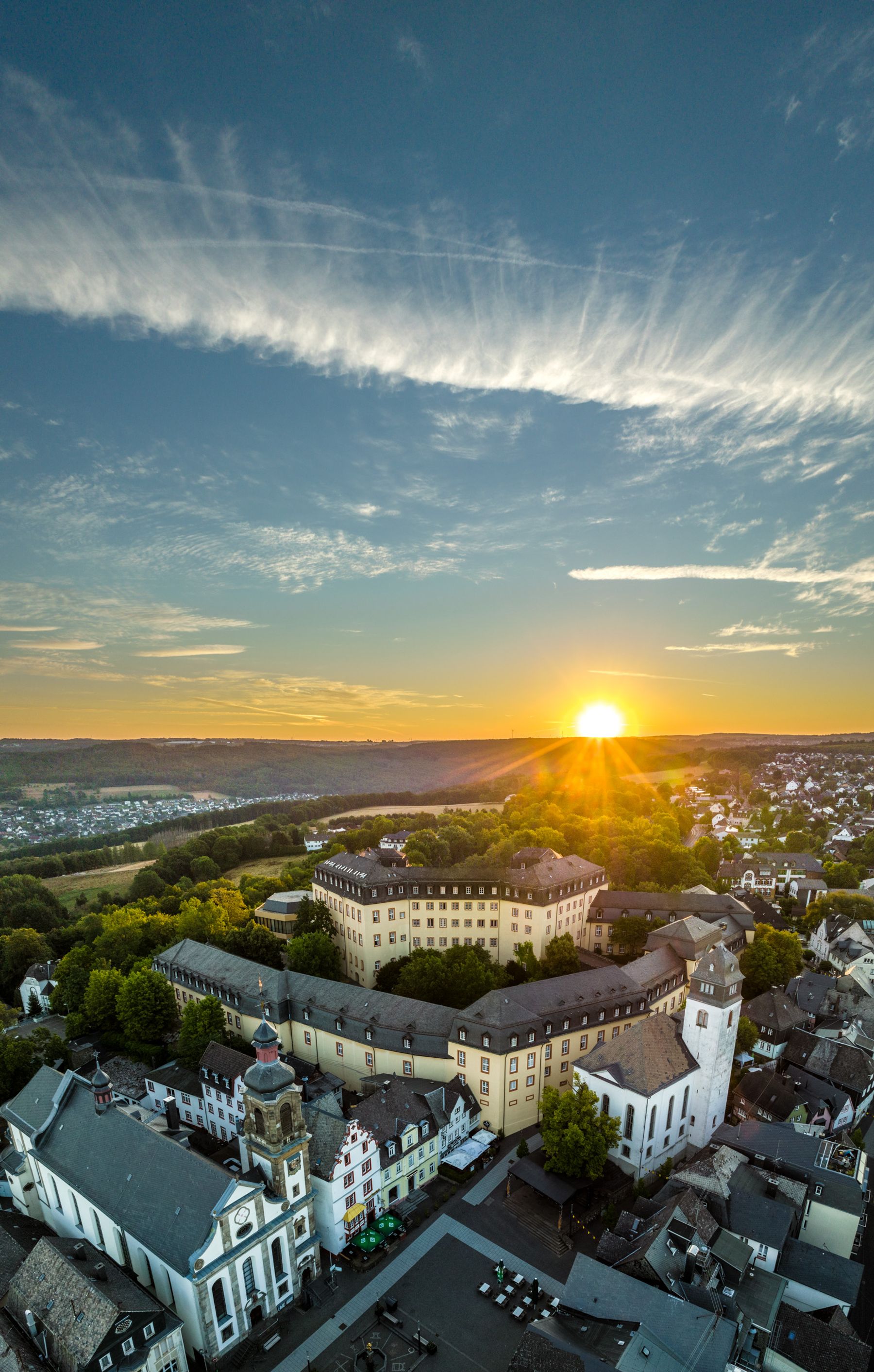Luftaufnahme einer historischen europäischen Stadt bei Sonnenuntergang mit einem großen schlossähnlichen Gebäude, das von Bäumen umgeben ist, während die Sonne über fernen Hügeln untergeht und der Himmel mit Wolkenfetzen gefüllt ist.